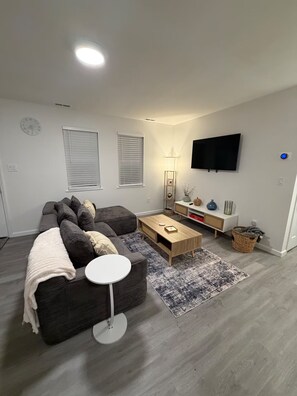 Un saló modern amb un sofà gris, una taula de centre, una televisió i una estant penjada a la paret amb objectes decoratius.