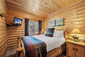 Een slaapkamer in een blokhut met een bed, een wandtelevisie, een raam met gordijnen en twee schilderijen aan de muur.