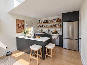 Eine moderne Küche mit einer zentralen Kochinsel, Barhocker, einem Kühlschrank und vielfältigem Küchenzubehör in Regalen.
