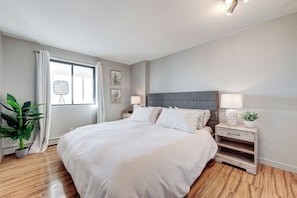 A modern bedroom with a large bed, bedside tables, a potted plant, and artwork on the walls. - The Skyside | 27th Floor | Penthouse + City Views (Saskatoon)