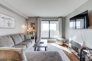 A modern living room with a grey sofa, a coffee table, a TV mounted on the wall, and a large window with curtains. - The Skyside | 27th Floor | Penthouse + City Views (Saskatoon)
