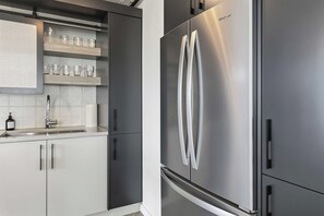 A modern kitchen with a stainless steel refrigerator, white cabinets, and a built-in shelf with glassware. - THE COSMOPOLITAN- YXE Penthouse (Saskatoon)
