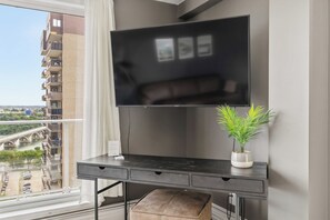 A modern living space with a flat-screen TV mounted on a dark console table, a small potted plant, and a view of a bridge and greenery outside. - THE COSMOPOLITAN- YXE Penthouse (Saskatoon)
