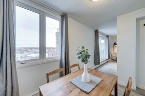 A dining area with a wooden table, chairs, a white vase with green plants, and a view of a cityscape through large windows. - The Bruno - 1 BR - Close to RUH (Saskatoon)