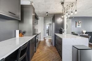 A modern kitchen with a marble countertop, stainless steel appliances, and a herringbone wood floor. - The Concord - Modern Condo Downtown YXE (Saskatoon)