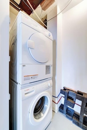 A stacked washer and dryer unit in a laundry room with a wooden rack and a white wall. - The Concord - Modern Condo Downtown YXE (Saskatoon)