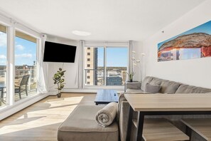 A modern living room with a sofa, a dining table, and a TV. There are large windows with a view of buildings and a cityscape. - THE BRADSHAW - 18th-floor 2 Bed Downtown Condo (Saskatoon)