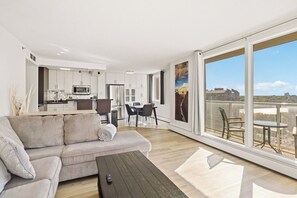 A modern living space with a sofa, dining table, and chairs, a kitchen area, and large windows offering a view of the outdoors. - THE BRADSHAW - 18th-floor 2 Bed Downtown Condo (Saskatoon)