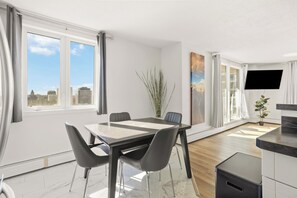 A modern dining area with a table and chairs, a potted plant, and a view of the city through large windows. - THE BRADSHAW - 18th-floor 2 Bed Downtown Condo (Saskatoon)