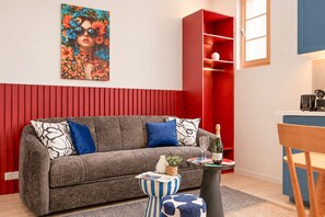 Ein Wohnzimmer mit grauem Sofa, rotem Regal und einem kleinen runden Tisch mit Flasche und Gläsern.