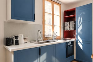 Eine kompakte Küche mit blauen Schränken, einer weißen Arbeitsplatte, einem Einbauofen und einem Fenster mit Holzjalousien.