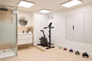 Ein modernes Badezimmer mit Duschkabine aus Glas, einem runden Spiegel, einer Wandtoilette und einer kleinen Ecke zum Trainieren mit Kurzhanteln.