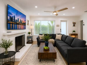 A living room with a fireplace, a large flat-screen TV, a modern sofa, a wooden coffee table, and a ceiling fan. - Your Perfect Fort Lauderdale Stay I 4BR, Private Pool & Free Parking (Fort Lauderdale)