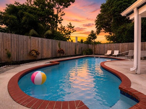 Una piscina con valla de madera, borde de ladrillo y atardecer de fondo.