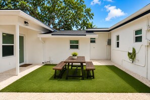A patio with a wooden table and benches, surrounded by white walls and a green lawn. - Your Perfect Fort Lauderdale Stay I 4BR, Private Pool & Free Parking (Fort Lauderdale)