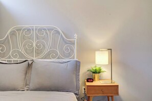 A bed with a white metal headboard, a bedside table with a lamp and a clock, and a small potted plant.
