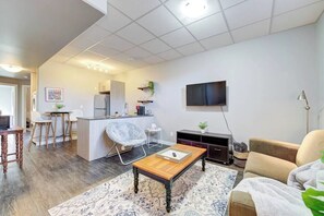 A modern living room with a wooden coffee table, a sofa, a TV mounted on the wall, and a kitchen area in the background. - The Arlo - North Park Gem Close to Spadina! (Saskatoon)