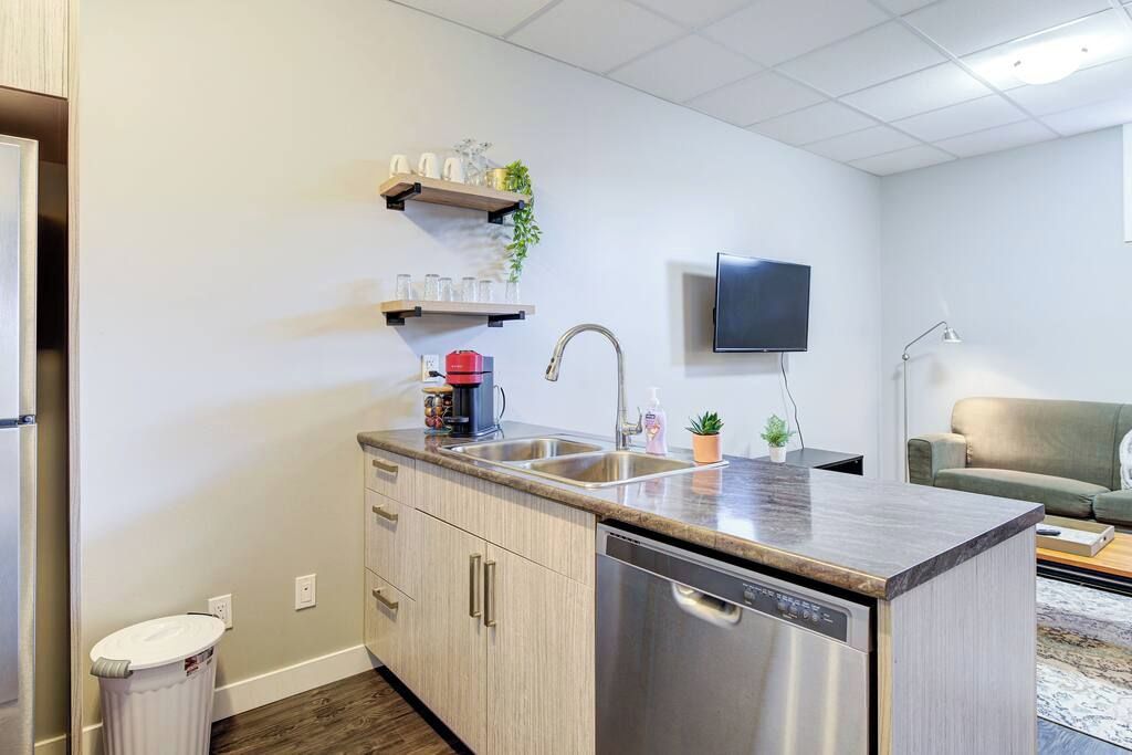 A compact kitchen with a dishwasher, a sink, and a countertop. There are cabinets, a trash can, and a television mounted on the wall.