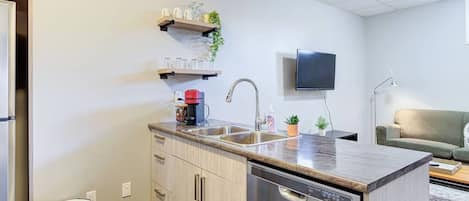A compact kitchen with a dishwasher, a sink, and a countertop. There are cabinets, a trash can, and a television mounted on the wall.