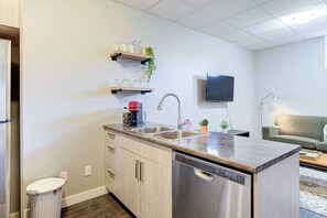 A compact kitchen with a dishwasher, a sink, and a countertop. There are cabinets, a trash can, and a television mounted on the wall. - The Arlo - North Park Gem Close to Spadina! (Saskatoon)