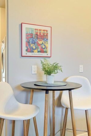Una habitación con una mesa redonda, dos sillas blancas, una obra de arte enmarcada en la pared y una planta en maceta sobre la mesa.