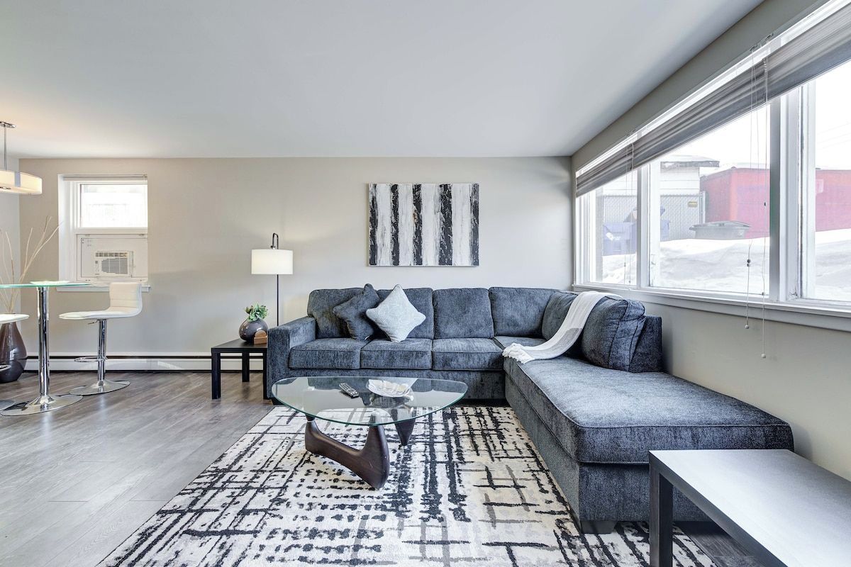 A modern living room with a sectional sofa, a glass coffee table, and a view of the outside through large windows.