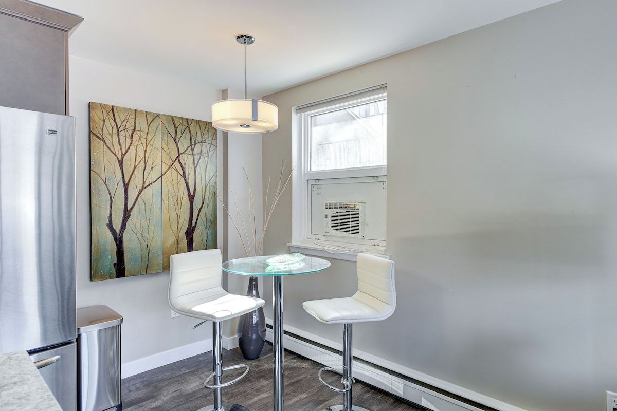 A modern kitchen with a refrigerator, a glass table with two chairs, and a painting on the wall.