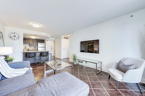 A modern living room with a sofa, armchair, and coffee table. A kitchen with a dining area is visible in the background. - The Tuscany - Located Downtown! (Saskatoon)