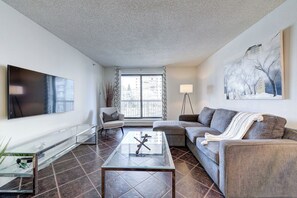 A modern living room with a flat-screen TV, a glass coffee table, a grey sofa, a white armchair, and a painting on the wall. - The Tuscany - Located Downtown! (Saskatoon)