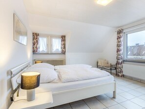 A bedroom with a bed, a bedside table with a lamp, a radiator, a window with curtains, and a view of buildings outside. - Holiday Apartment With 1 Bedroom (St. Peter-Ording)