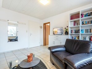 A modern living room with a leather sofa, a bookshelf, a television, and a dining area with a table and chairs. - Holiday Apartment With 1 Bedroom (St. Peter-Ording)