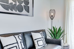 Ein modernes Wohnzimmer mit einem schwarzen Ledersofa, zwei gemusterten Zierkissen, einer Stehlampe und einer großen Grünpflanze.
