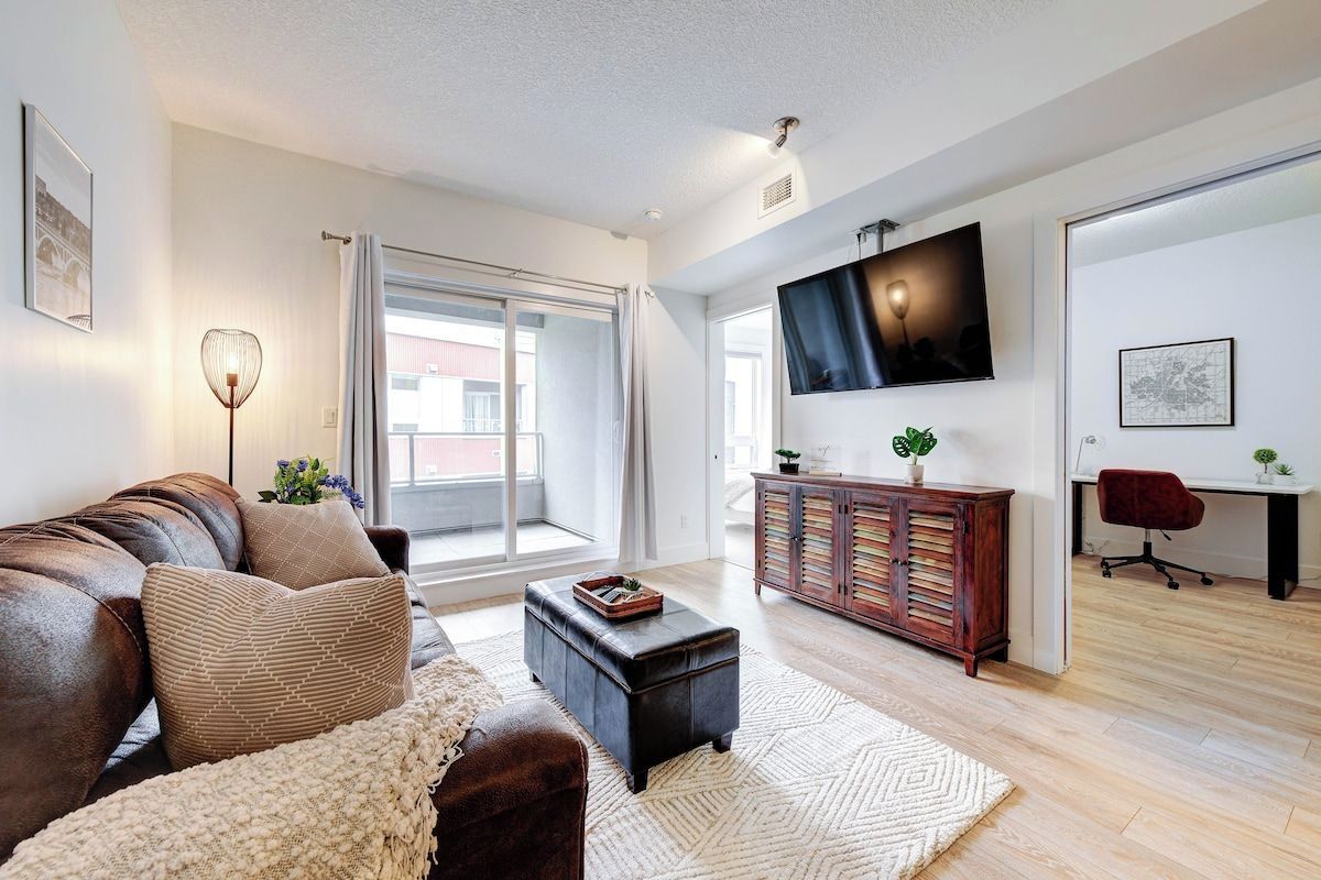 A modern living room with a leather sofa, a coffee table, a TV mounted on the wall, and a sliding glass door leading to a balcony.