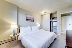 Ein Hotelzimmer mit einem Bett, Nachttischlampen, einer Uhr, einem Gemälde an der Wand und einem Kleiderschrank mit Regalen.