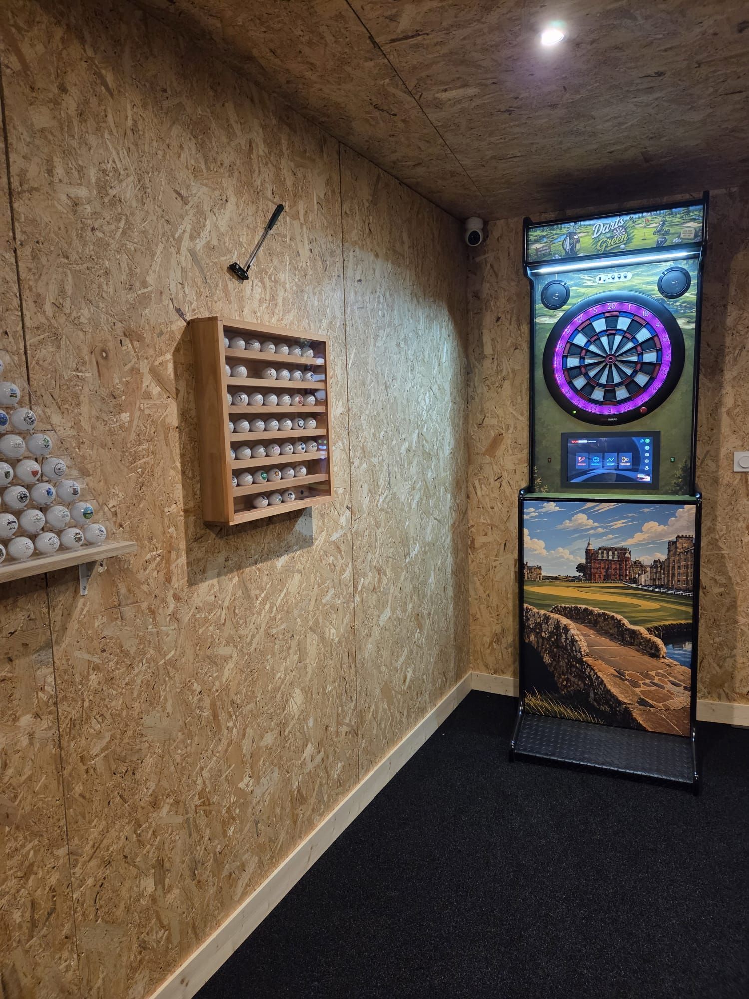 A room with a dartboard, a shelf with darts, and a digital screen displaying a game.