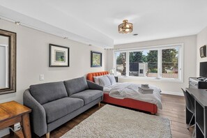A modern bedroom with a grey sofa, a bed with an orange headboard, a desk, and a large window with a view of the outdoors. - The Nest - Cozy 1BR | Free Parking (Saskatoon)