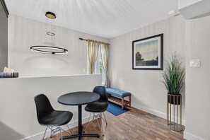Ein modernes Zimmer mit einem runden Tisch, zwei Stühlen, einer Bank, einer Pendelleuchte, einem gerahmten Bild an der Wand und einer hohen Pflanze in einem goldenen Topf.