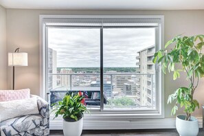 A balcony with a view of a cityscape, a sofa, a lamp, and two potted plants. - The Oslo - 2BD Downtown (Saskatoon)