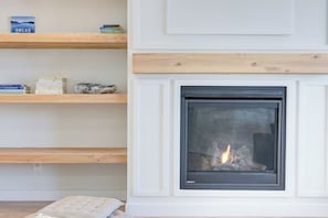 A fireplace with a visible fire, wooden shelves with books, and a cushion on the floor. - Hygge Haven – Stylish Stay near the River (Saskatoon)