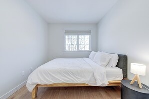 A bedroom with a bed, a window with blinds, a bedside lamp, and a wooden floor. - Hygge Haven – Stylish Stay near the River (Saskatoon)