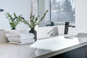 A bathroom with a white countertop, a black vase with green plants, and a stack of white towels. - Hygge Haven – Stylish Stay near the River (Saskatoon)