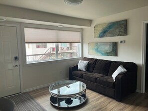 A living room with a brown sofa, a glass coffee table, a door, and a window with blinds. - The Magnolia - 2BDR Condo Downtown (Saskatoon)
