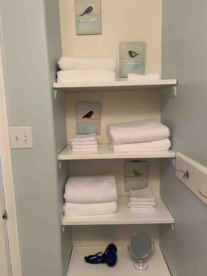 A bathroom with white shelves holding folded towels, a wall-mounted light switch, and a small mirror. - The Magnolia - 2BDR Condo Downtown (Saskatoon)