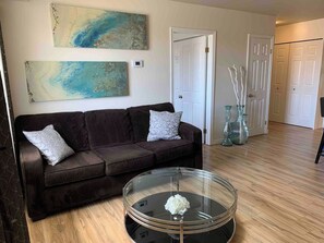 A living room with a dark brown sofa, two patterned pillows, a glass coffee table with a white flower, and abstract wall art. - The Magnolia - 2BDR Condo Downtown (Saskatoon)