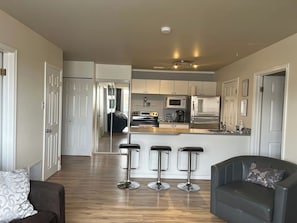 A modern kitchen with a central island, bar stools, and a seating area with a sofa and armchair. - The Magnolia - 2BDR Condo Downtown (Saskatoon)