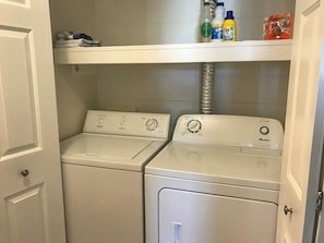 A laundry room with two white washing machines and a shelf above containing cleaning supplies. - The Magnolia - 2BDR Condo Downtown (Saskatoon)