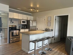 A modern kitchen with stainless steel appliances, a central island with bar stools, and a wooden floor. - The Magnolia - 2BDR Condo Downtown (Saskatoon)