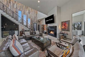 A spacious living area with a fireplace, leather sofas, a coffee table, and a staircase with a wooden wall. - Lake Buchanan Bliss | Pool & Spa • Lakefront Beach (Burnet)