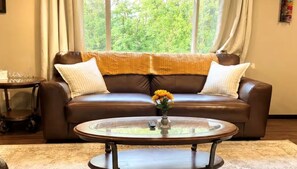 A brown leather sofa with a yellow throw blanket and white pillows, a glass coffee table with a vase of flowers, and a view of greenery through large windows. - Luxury 6BR Retreat - Spacious & Refined Living (Apple Valley)
