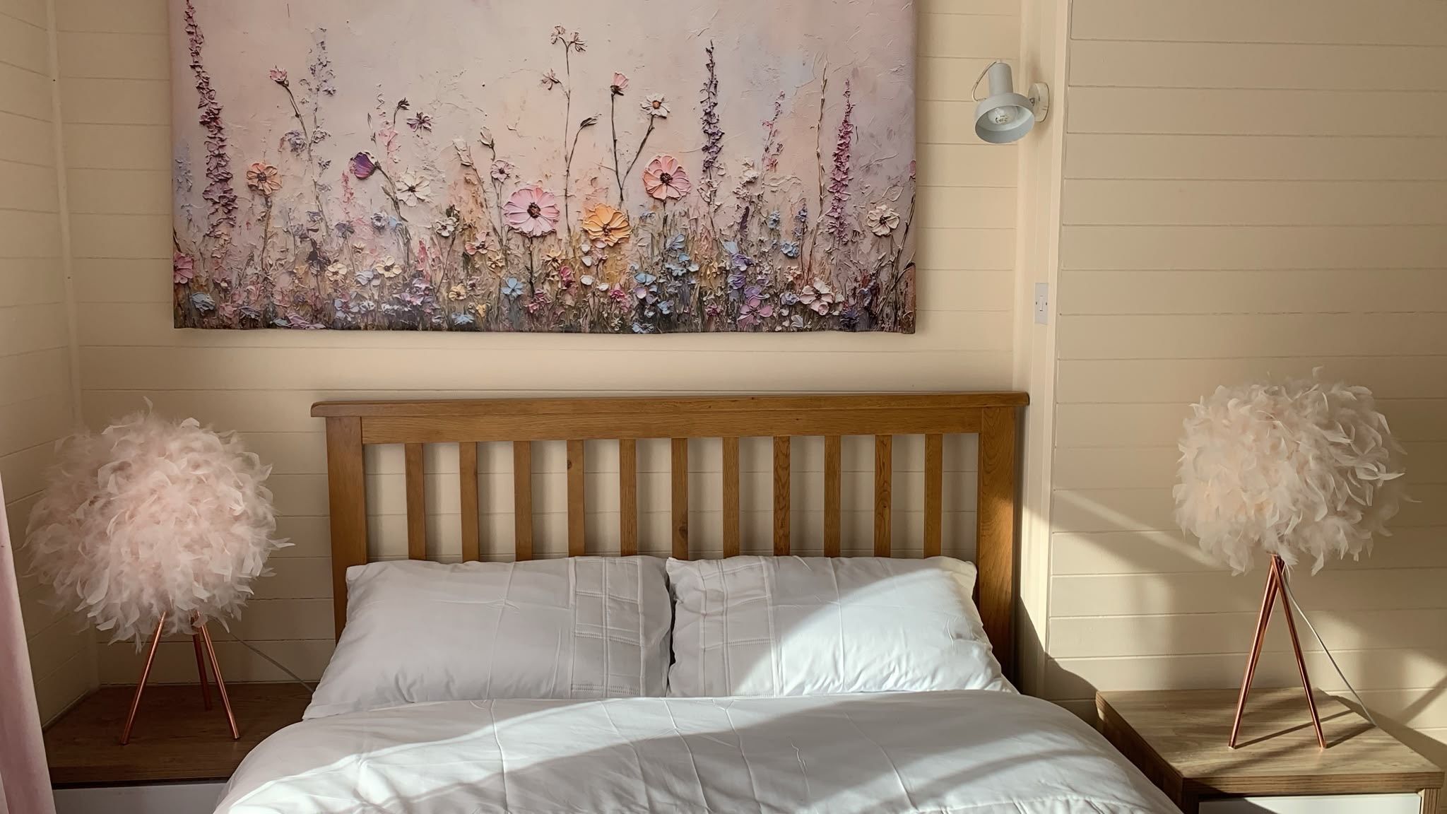 Un dormitorio con cabecera de madera, ropa de cama blanca, una obra de arte floral en la pared y dos mesitas de noche con adornos blancos y suaves.